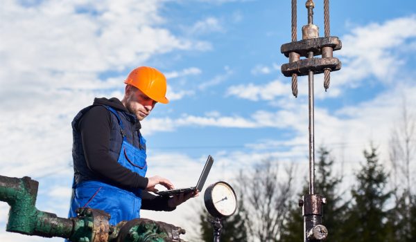 Engineer in work overalls and orange helmet checking oil pumping unit at oil field, using laptop. Male worker standing in front of oil well pump jack. Concept of petroleum industry, oil extraction.